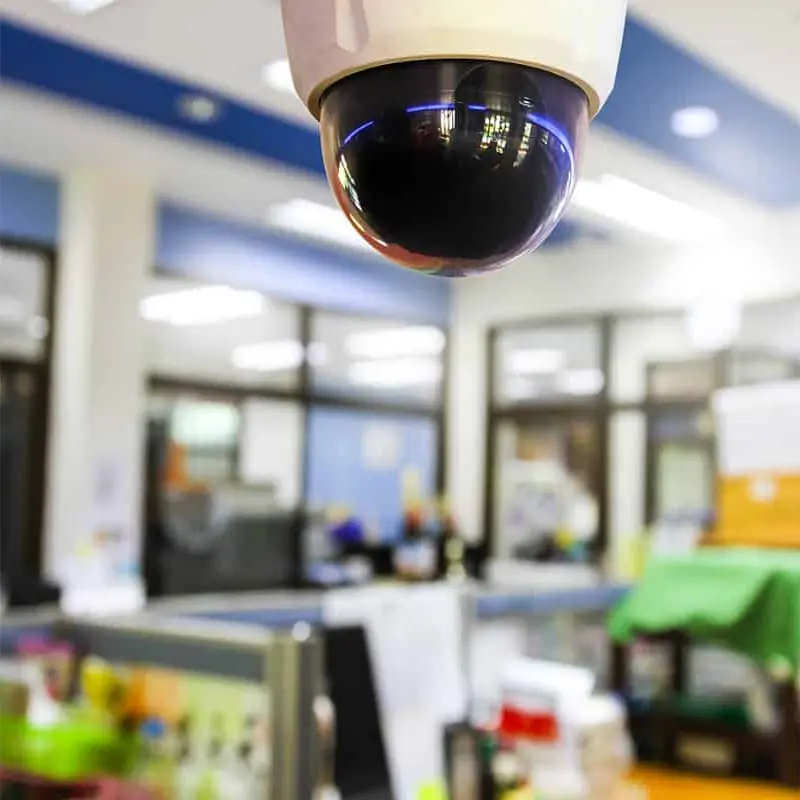 Image of a security camera hanging from the ceiling of an office building. The camera is pointed at the main entrance of the building and is used to monitor the employees and customers of the building.